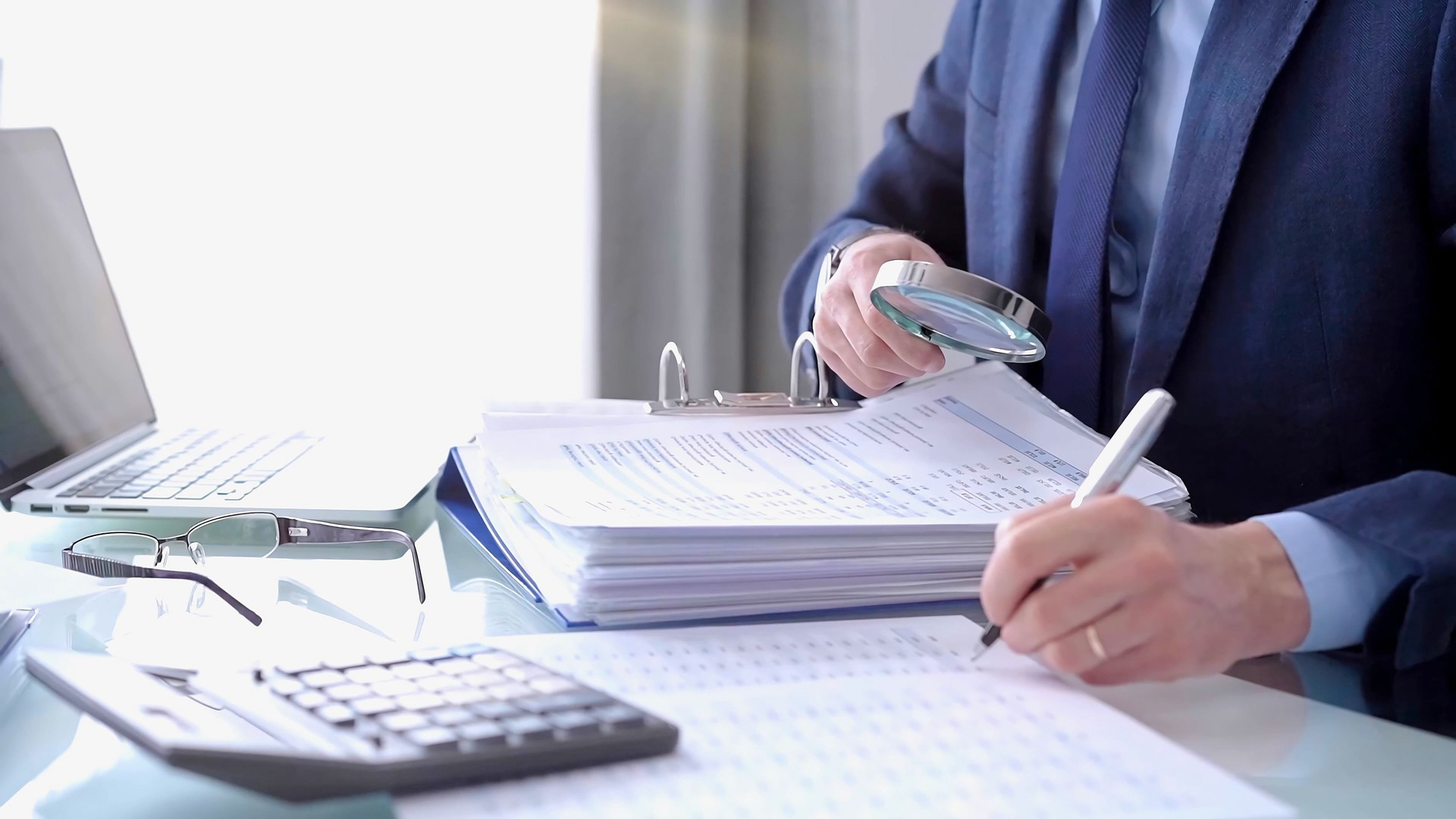 Businessman using a calculator and magnifying glass while analyzing financial documents at desk in fair modern office. Audit and taxes in business